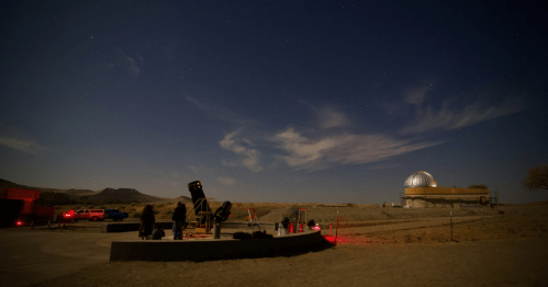 A night scene at an observatory with telescopes, people observing the sky, and a dome structure under a starry sky.