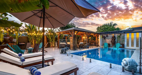 A serene backyard pool area with lounge chairs, an umbrella, and tropical plants, set against a colorful sunset sky.
