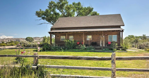 A rustic log cabin surrounded by greenery and a wooden fence under a clear blue sky.