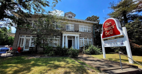 A charming brick building with a sign reading "The Dinner Bell," surrounded by greenery and a clear blue sky.