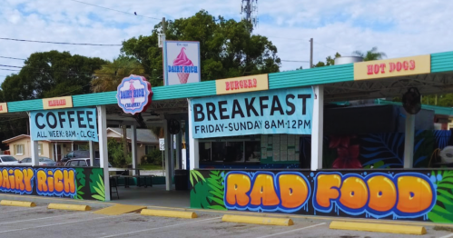 Colorful food stand with signs for coffee, breakfast, burgers, and hot dogs, featuring vibrant murals.