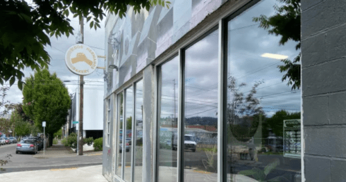 A bakery storefront with large windows, a sign, and trees lining the street under a cloudy sky.
