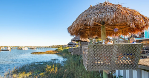 A scenic view of thatched-roof huts by a calm waterway, with people enjoying the outdoors on a sunny day.