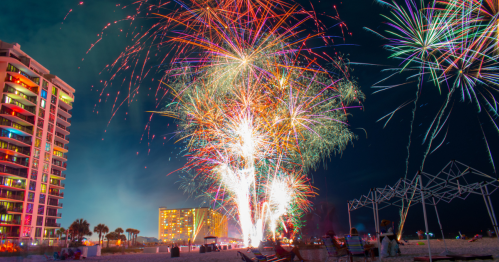 Colorful fireworks illuminate the night sky over a beach, with buildings and palm trees in the background.