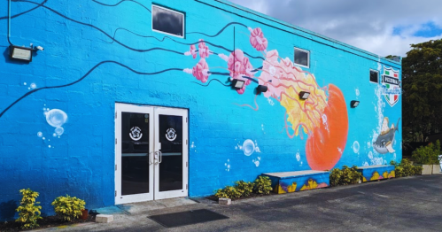 Vibrant blue mural on a building featuring jellyfish, bubbles, and a shark, with double glass doors in front.