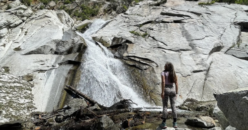 A person stands near a waterfall cascading over rocky terrain in a natural setting.