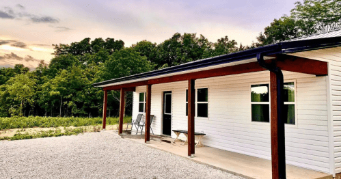 A modern white house with a porch, surrounded by trees and a gravel pathway, under a colorful sunset sky.