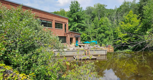 A brick building by a calm water body, surrounded by lush greenery and outdoor seating with umbrellas.