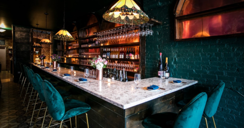A stylish bar with a marble countertop, green velvet stools, and shelves lined with wine bottles and glasses.