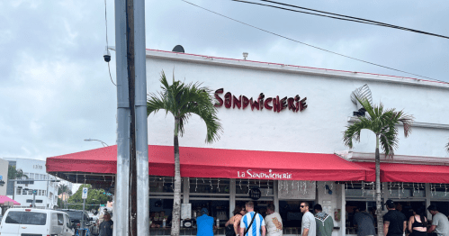 A busy sandwich shop with a red awning, palm trees, and people waiting outside on a cloudy day.
