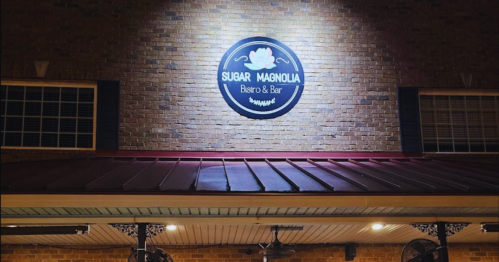 Sign for "Sugar Magnolia Bistro & Bar" illuminated on a brick wall, with a decorative logo above the entrance.