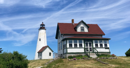 A white lighthouse beside a historic house, set against a blue sky with wispy clouds. Green grass surrounds the scene.