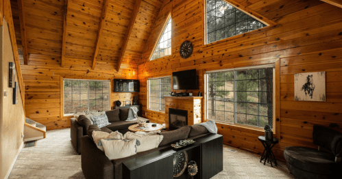 Cozy wooden cabin interior with large windows, a comfortable sofa, and a TV, surrounded by nature.