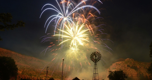 Colorful fireworks burst in the night sky above a silhouette of a windmill and mountains.