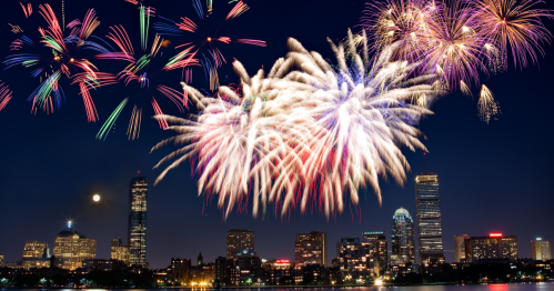 Colorful fireworks explode over a city skyline at night, with a full moon visible in the background.