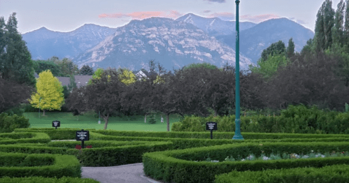 A scenic garden with neatly trimmed hedges, surrounded by mountains and trees under a pastel sky at dusk.