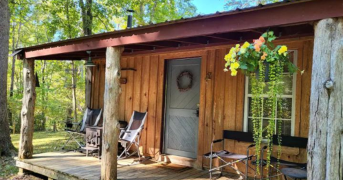 A rustic wooden cabin with a porch, featuring two chairs and hanging flowers, surrounded by trees.