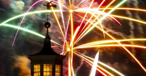 Colorful fireworks burst in the night sky above a silhouetted building with a weather vane.