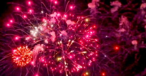 Colorful fireworks explode in a night sky, creating vibrant bursts of pink, white, and gold against a dark background.