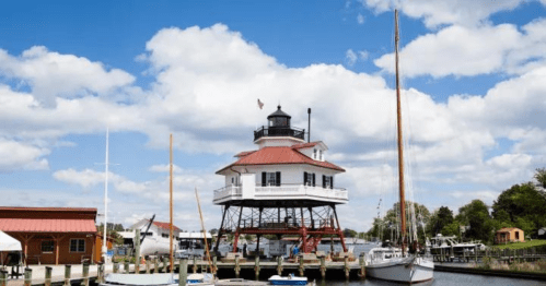 A lighthouse on stilts by a marina, surrounded by boats and under a blue sky with fluffy clouds.