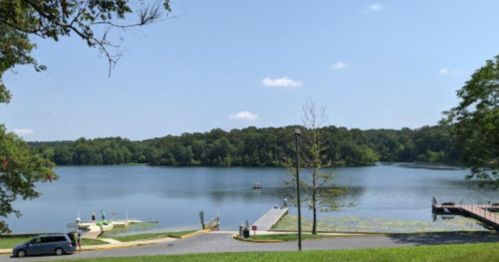 A serene lake surrounded by trees, with a boat ramp and dock visible in the foreground under a clear blue sky.