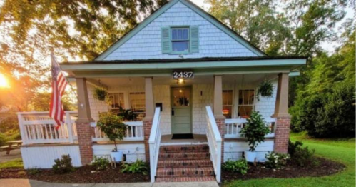 A charming white house with a porch, American flag, and lush greenery, bathed in warm sunset light.