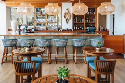 A modern bar with wooden furniture, blue bar stools, and pendant lights, featuring a well-stocked shelf behind the counter.