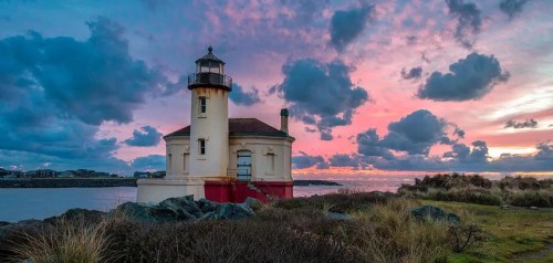 A lighthouse stands by the water under a colorful sunset, with clouds scattered across the sky and rocky foreground.