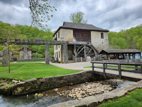 Historic stone mill with a waterwheel, surrounded by greenery and a small stream, under a cloudy sky.