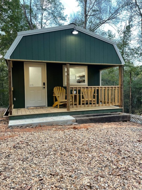 A small green cabin with a porch, featuring wooden chairs and a table, surrounded by trees and gravel.