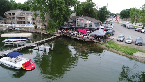 A waterfront restaurant with outdoor seating, boats docked, and a crowd enjoying the view on a sunny day.