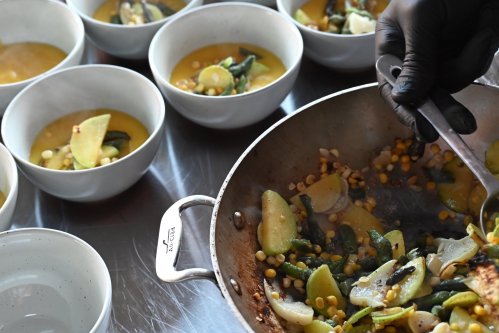 A chef in black gloves serves a vegetable dish from a pan into white bowls filled with corn and green vegetables.