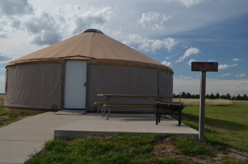 A tan yurt with a white door, picnic table, and fire pit, set against a cloudy sky and grassy landscape.