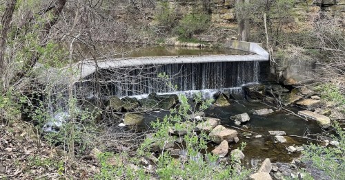 A small waterfall cascades over a concrete barrier into a rocky stream, surrounded by greenery and trees.