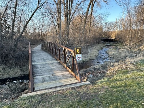 A wooden bridge over a small stream, surrounded by trees and grass, with a warning sign nearby.