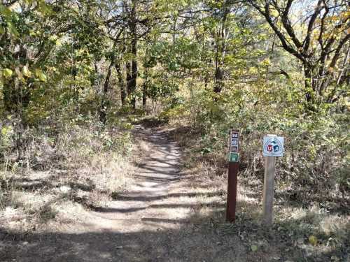 A narrow dirt path leads into a wooded area, marked by trail signs on either side.