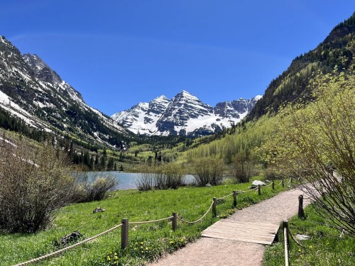 A scenic pathway leads to a lake surrounded by lush greenery and snow-capped mountains under a clear blue sky.