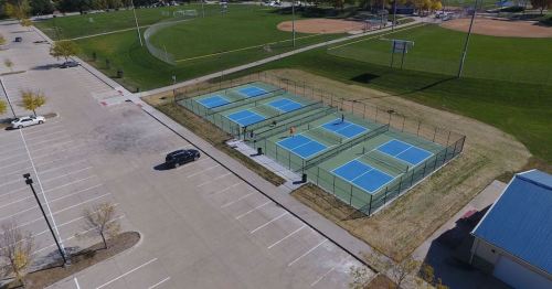Aerial view of tennis courts surrounded by a parking lot and sports fields in a park setting.