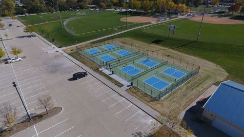 Aerial view of a sports complex featuring multiple tennis courts, a parking lot, and green fields in the background.