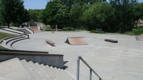 A sunny skate park featuring ramps, stairs, and green trees in the background.