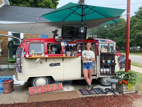 A woman stands by a vintage food truck, smiling, with a colorful awning and various items displayed around her.