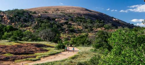 Two people walk along a dirt path towards a large, rocky hill surrounded by greenery under a blue sky.