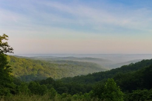 A serene landscape of rolling green hills under a blue sky, with mist gently covering the valleys.