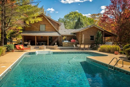 A serene backyard with a pool, surrounded by trees and a log cabin, featuring lounge chairs and a sunny sky.