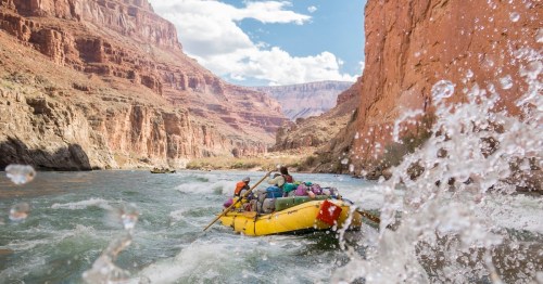 A group of people rafting on a river surrounded by red rock cliffs and splashing water under a blue sky.