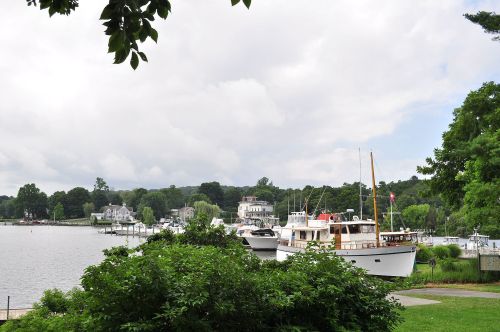 A serene waterfront scene with boats docked, lush greenery, and cloudy skies in the background.