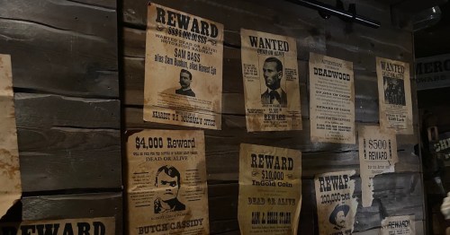 Old wanted posters on a wooden wall, featuring rewards for outlaws like Sam Bass and Butch Cassidy.