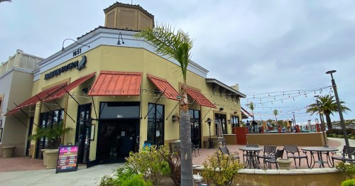 A restaurant with a tan exterior, red awnings, and outdoor seating, surrounded by palm trees and string lights.