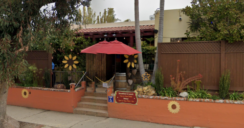 A charming restaurant entrance with a red umbrella, flower decorations, and a sign, surrounded by greenery.