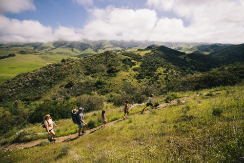 A group of hikers walking along a trail in a lush green landscape with rolling hills and cloudy skies.
